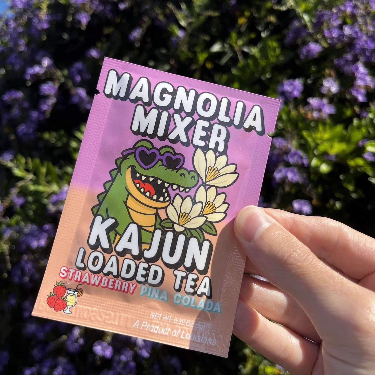 Hand holding a 'Magnolia Mixer Kajun Loaded Tea' packet with a colorful design against a floral background.